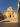 A wide-angle photograph of a historic beige stone church in Vicoria, Gozo facade on a hill, accessed by a long flight of steps under a clear blue sky. The image is framed by buildings on the right and left, with the church centered in the background.